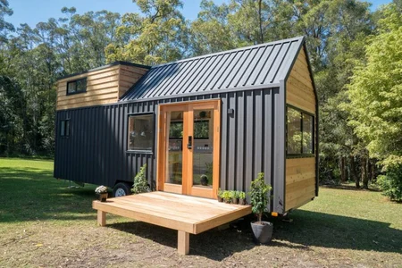 A modern tiny house on wheels with black metal siding and natural wood accents, featuring large windows, double glass doors, and a small wooden porch, serves as a stylish Accessory Dwelling Unit set in a grassy area surrounded by trees.